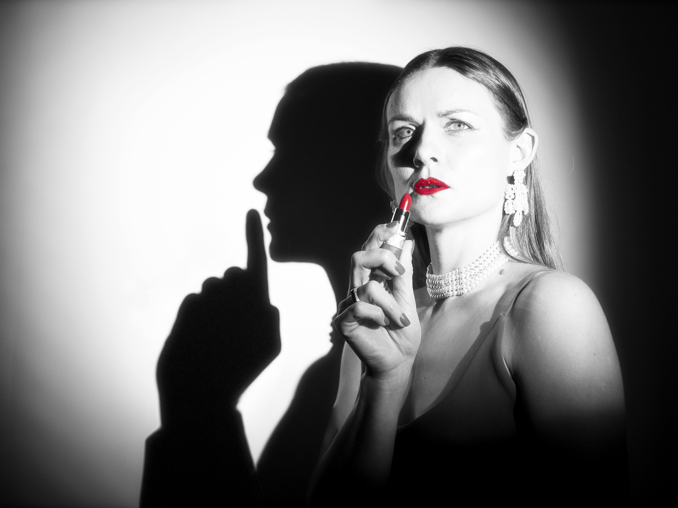 Production image from As SHE Likes It in black and white. A woman staring out of the image holding a bright red lipstick to her lips, the reflection looks like it's a gun